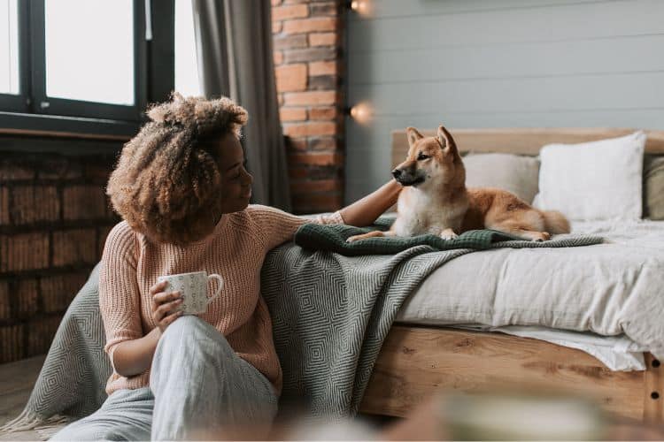 woman sitting at foot of bed, mug of beverage in hand, patting a dog