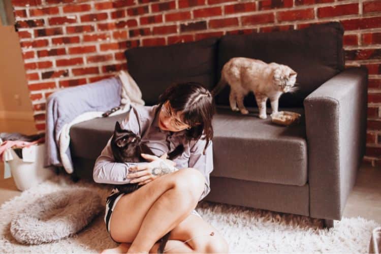 Woman sitting on floor beside a couch. She is cradling a cat in her arms. On the couch behind her is another cat