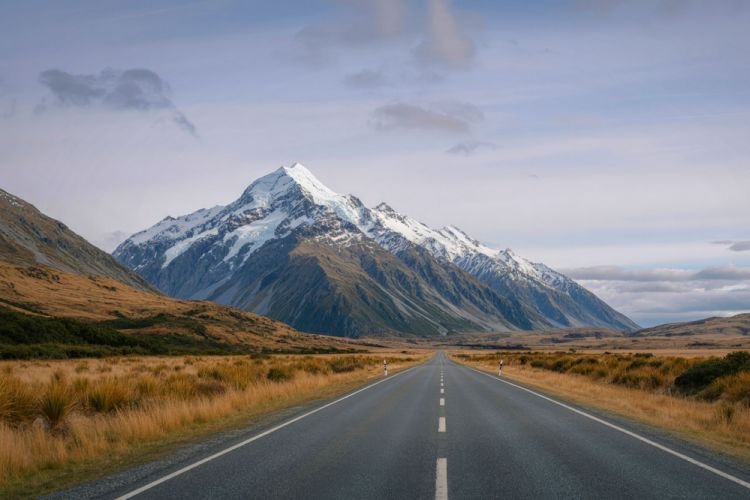 A long, straight road leading to a snow-covered craggy mountain in the distance