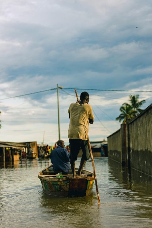 Man paddling a boat with a passenger, in a flooded village street